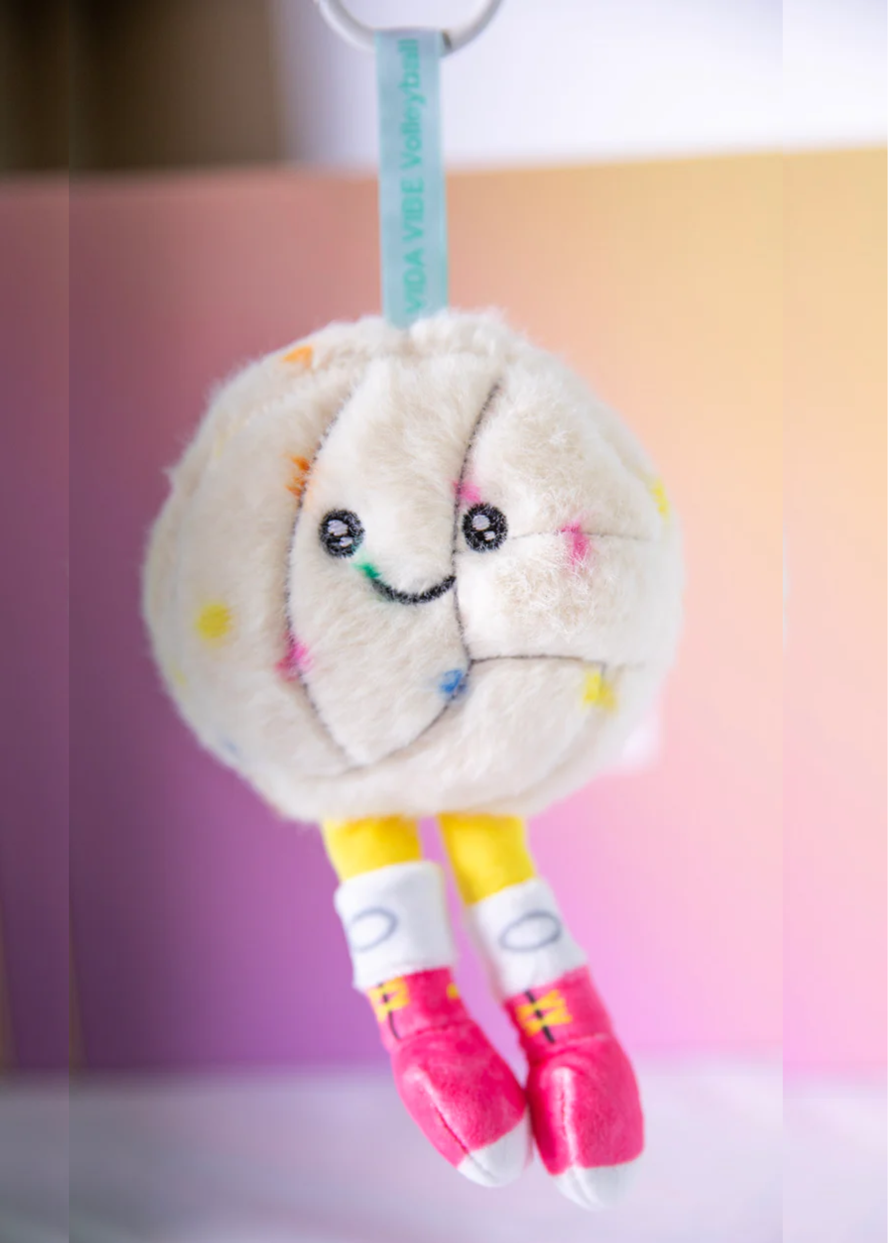 Plush volleyball keychain with smiling face, colorful polka dots, and pink shoes on a gradient background