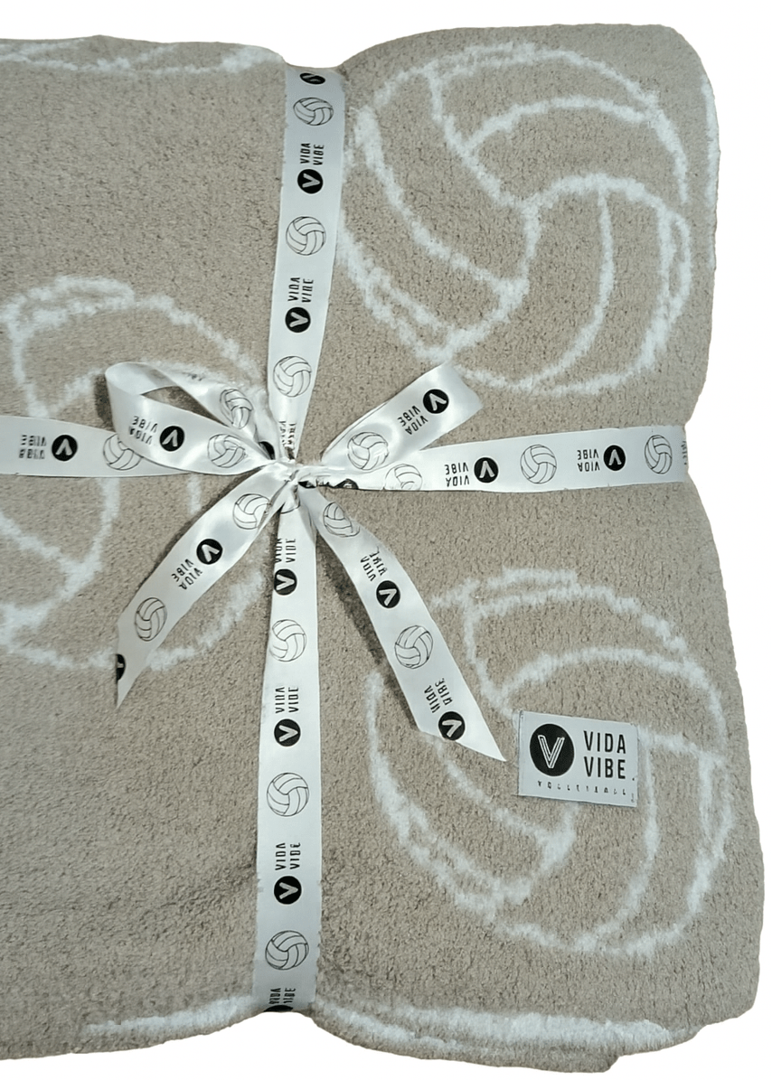 Folded beige blanket with white volleyball patterns and a ribbon, branded 'Vida Vibe'.