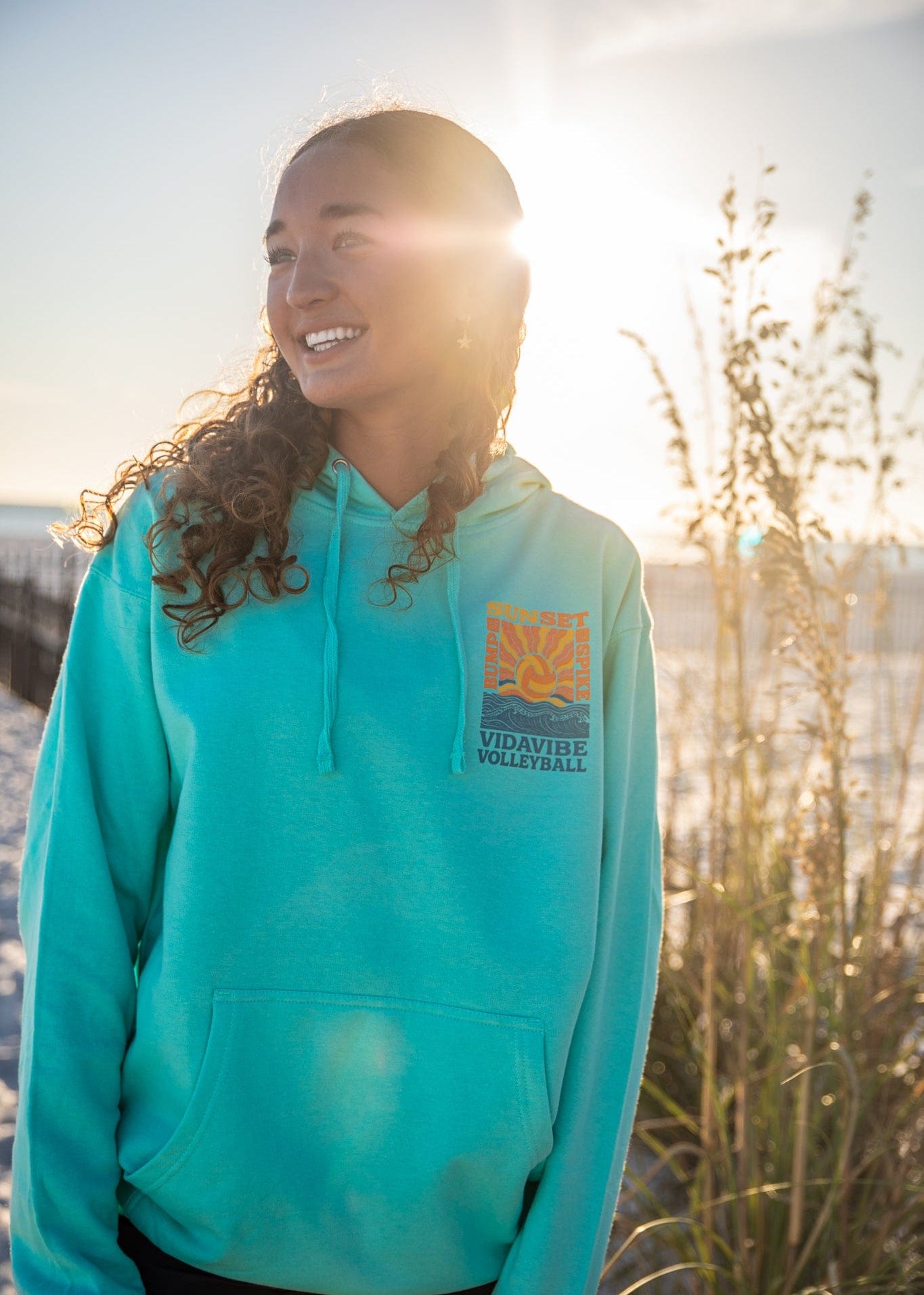 Person wearing a teal hoodie with a logo, standing outdoors with sunlight and natural background