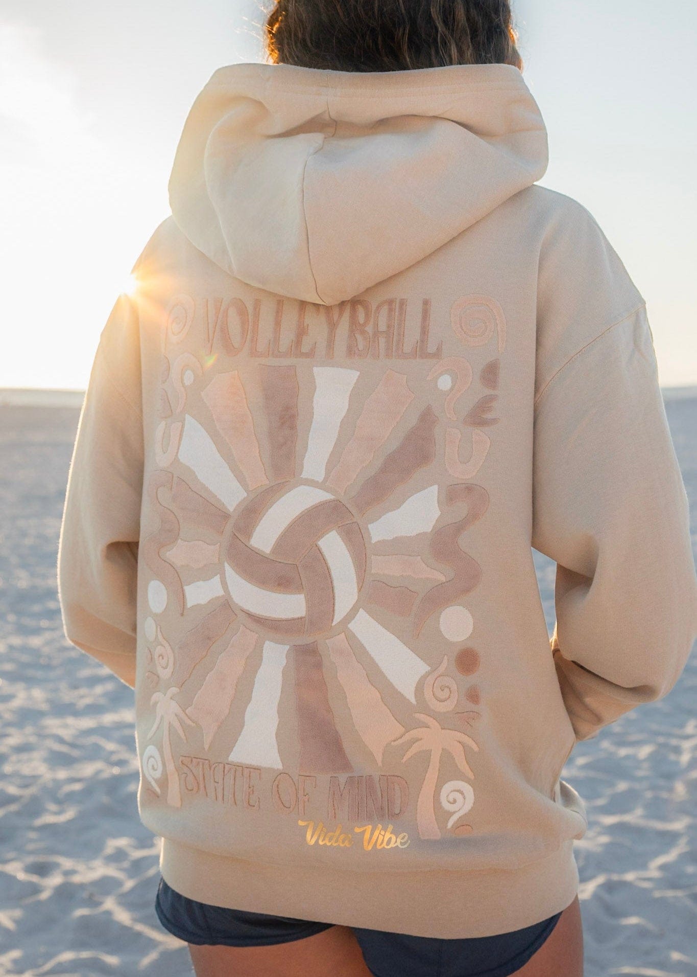 Person wearing a beige volleyball hoodie with volleyball design on a beach