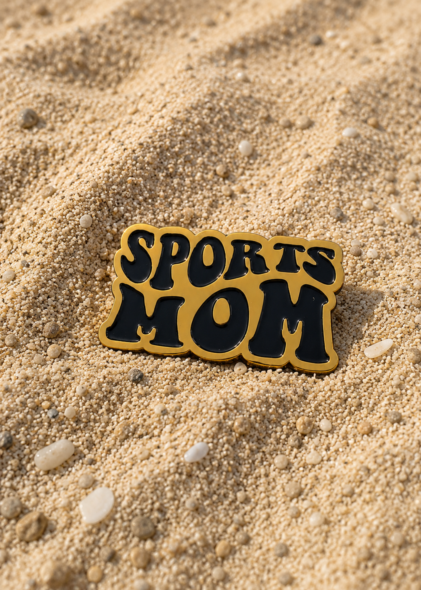 A gold and black enamel pin that reads 'SPORTS MOM' in bold retro lettering, resting on a sandy beach background.