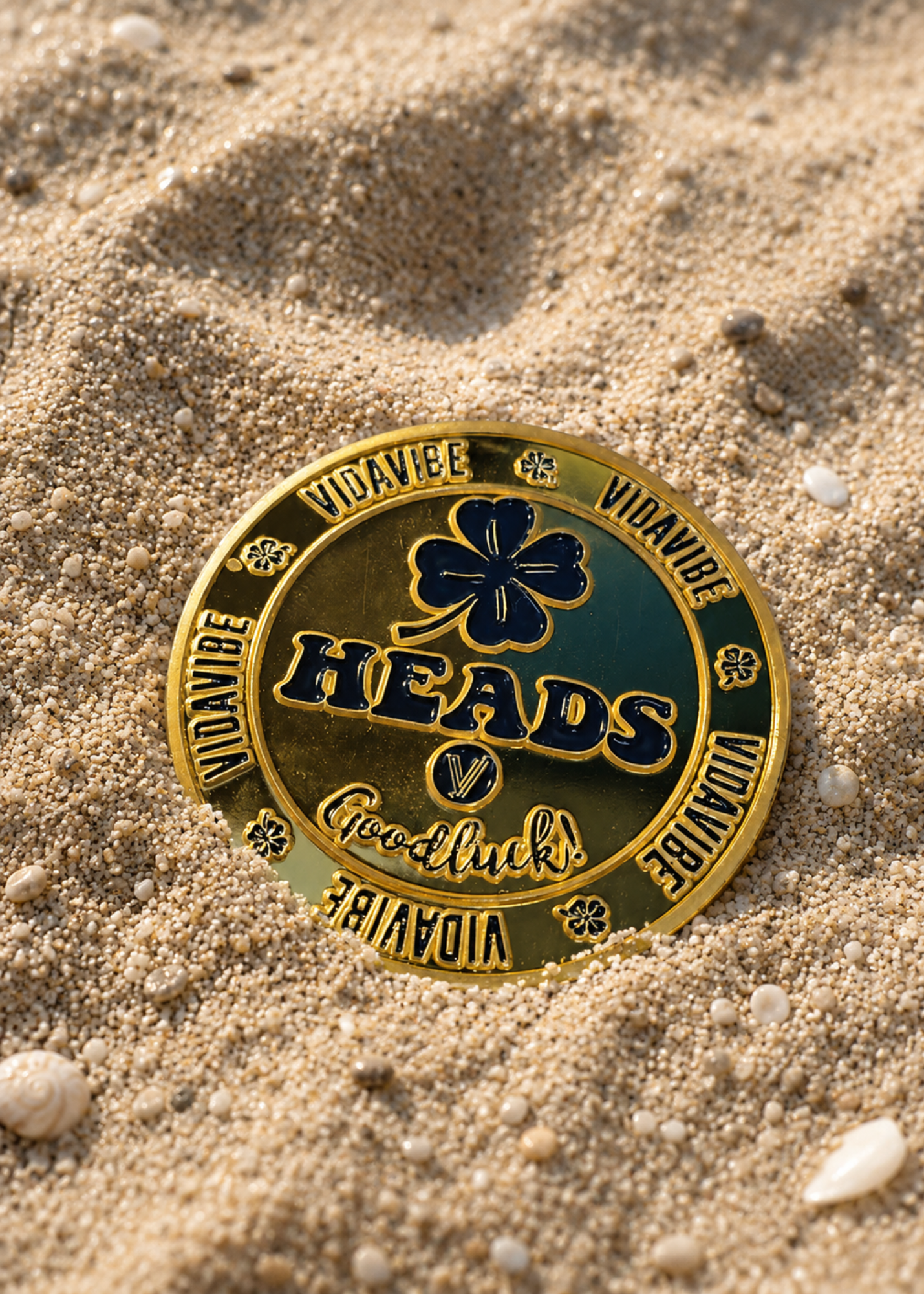 A circular gold and green challenge coin featuring a four-leaf clover and the word 'HEADS' with 'Vidavibe' around the edge, resting on sandy ground.