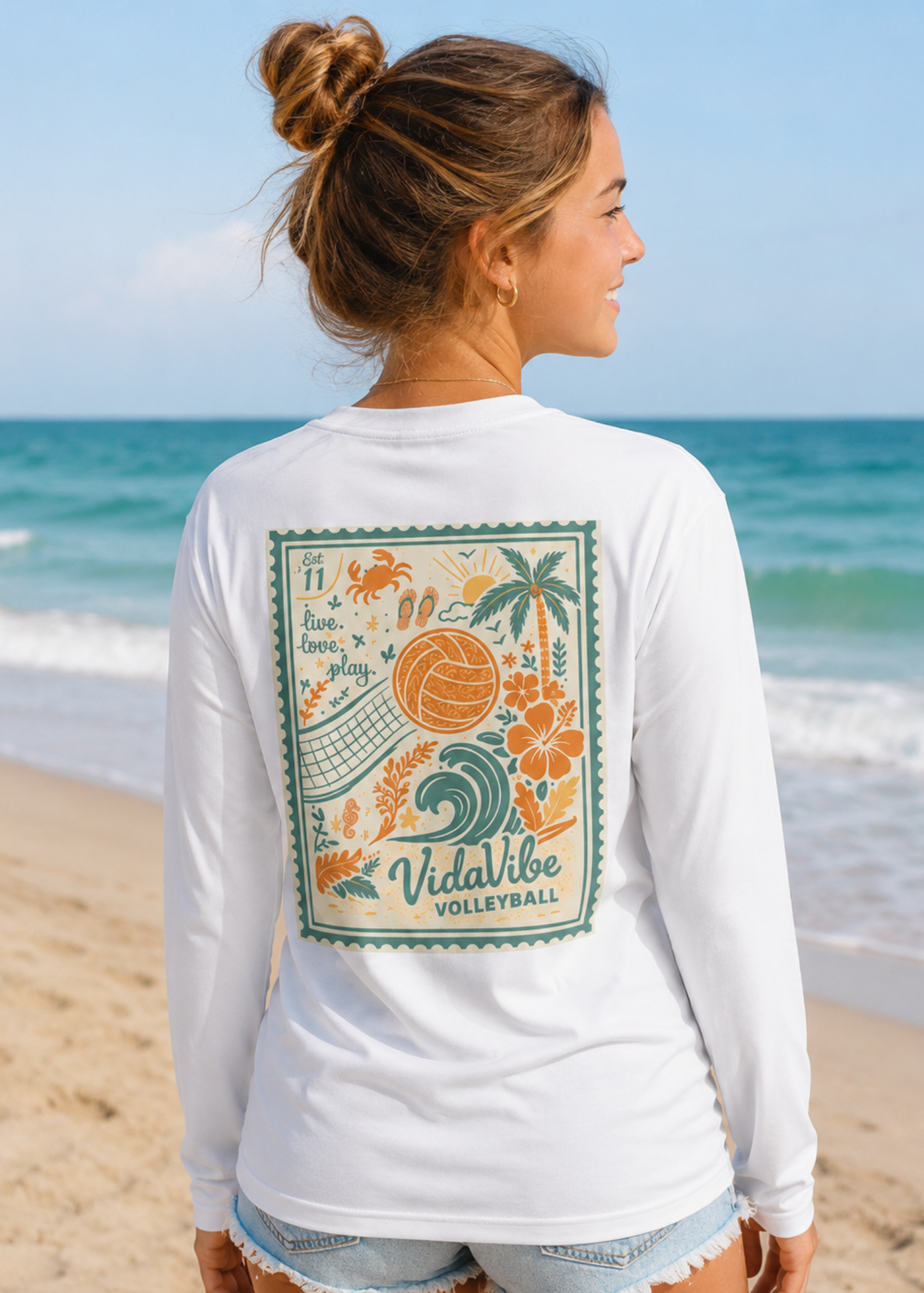 A woman standing on a sandy beach facing the ocean, wearing a white long-sleeve t-shirt with a colorful retro-style 'Vitalibe Volleyball' graphic printed on the back.