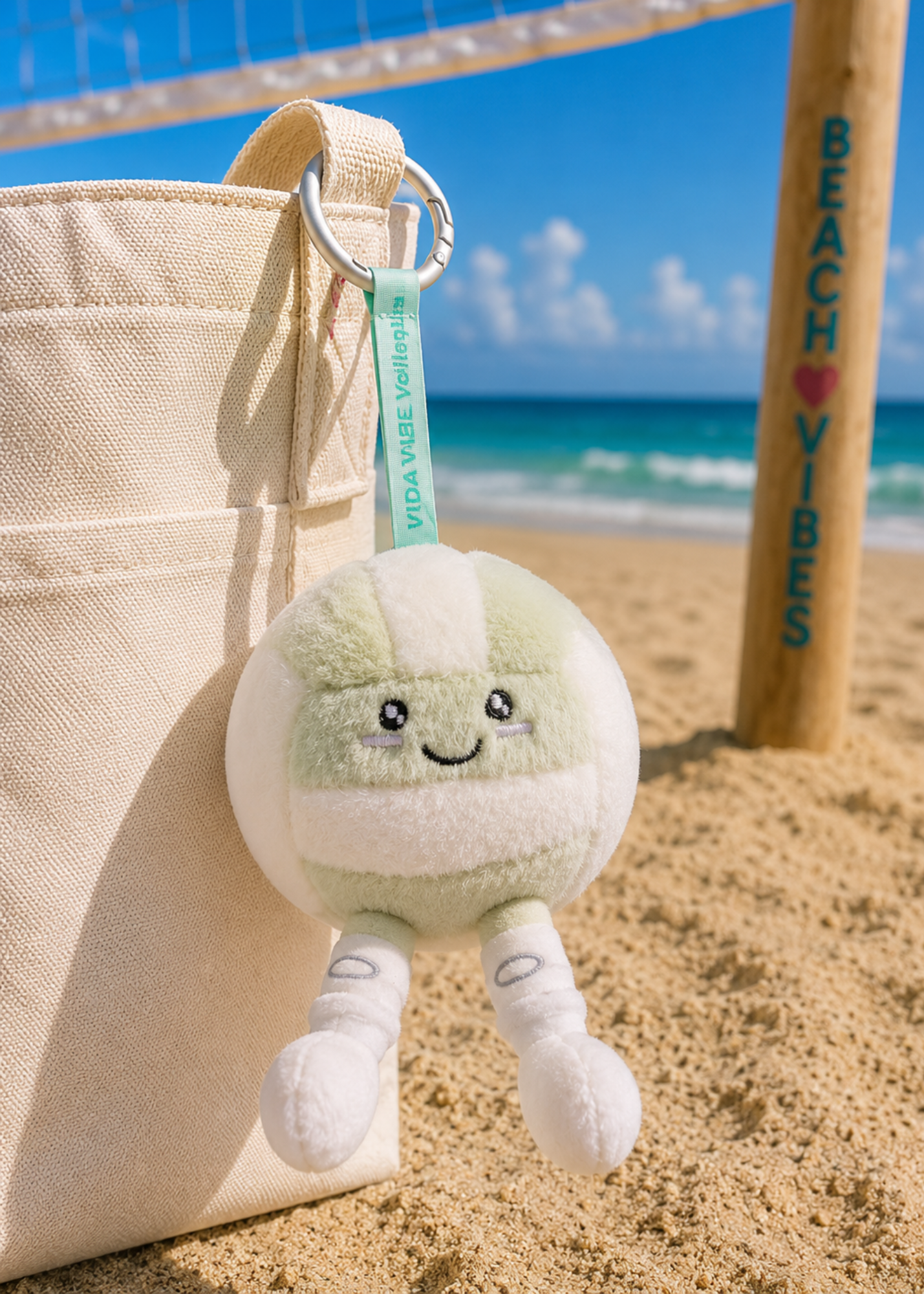 A plush volleyball character keychain hanging from a beige canvas tote bag on a sandy beach. The keychain features a cute smiling face and small legs, attached to a light green ribbon tag that reads 'VIDA VIBE VOLLEYBALL'.