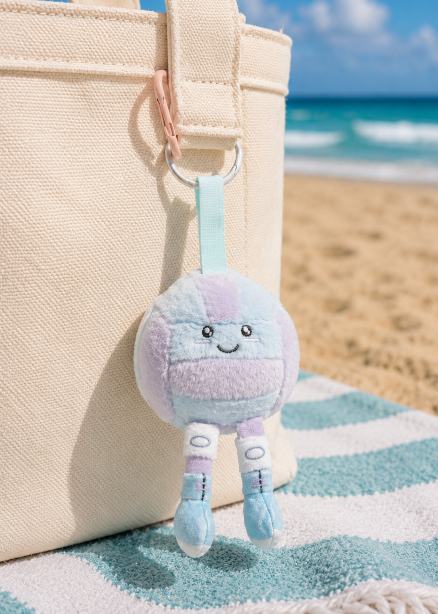 A pastel plush volleyball keychain character with a smiling face hangs from a cream canvas tote bag on a beach towel, with a sandy beach and ocean in the background.
