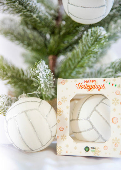 White volleyball-shaped ornaments with 'Happy Volleydays' packaging in front of a Christmas tree.