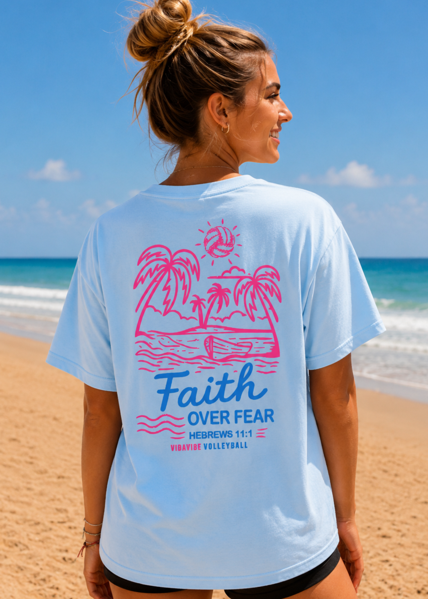 Woman on a beach wearing a light blue t-shirt with a pink palm tree design and 'Faith Over Fear' text.