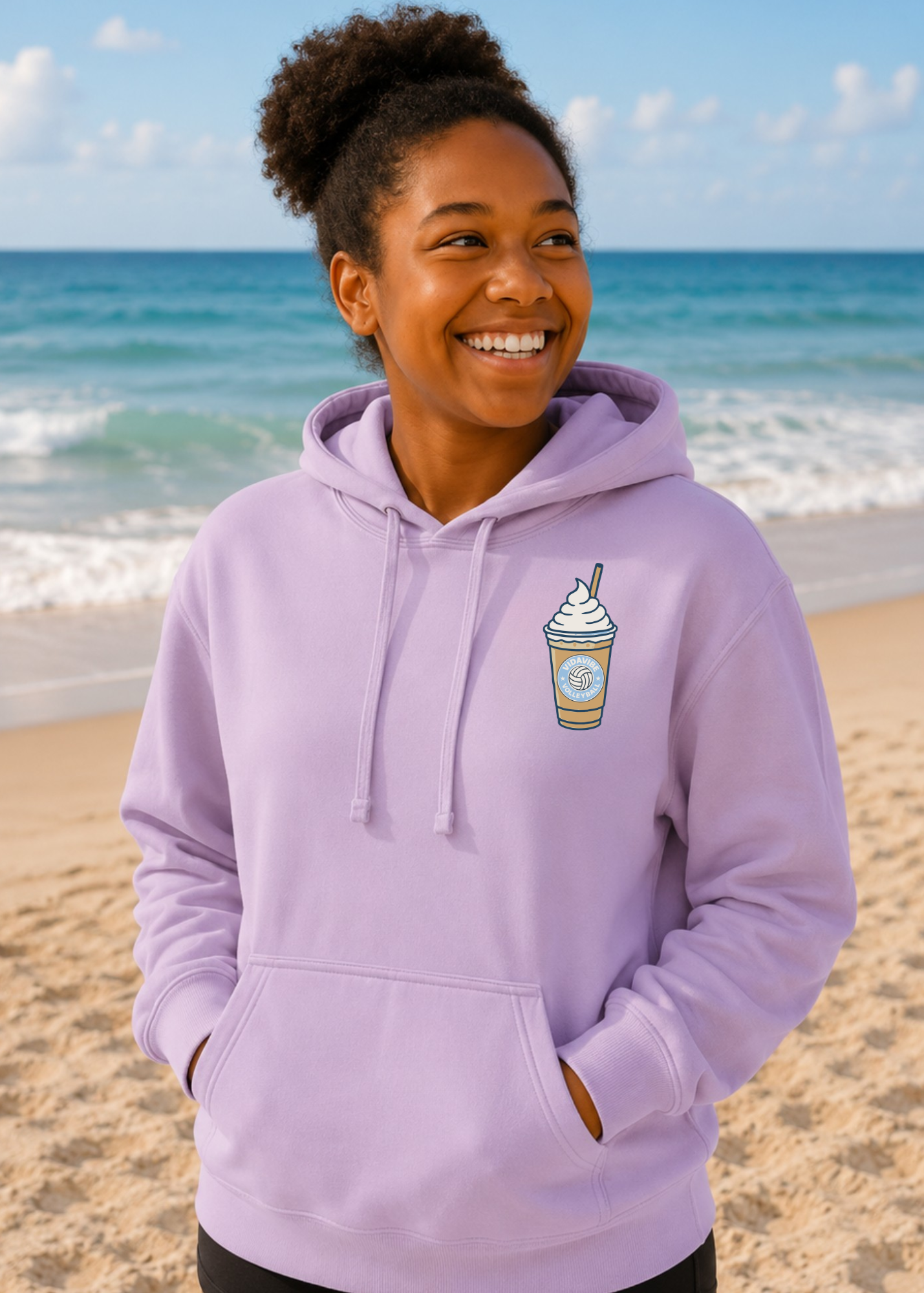 A woman smiling on a sandy beach wearing a light purple hoodie with a small coffee cup graphic on the left chest.