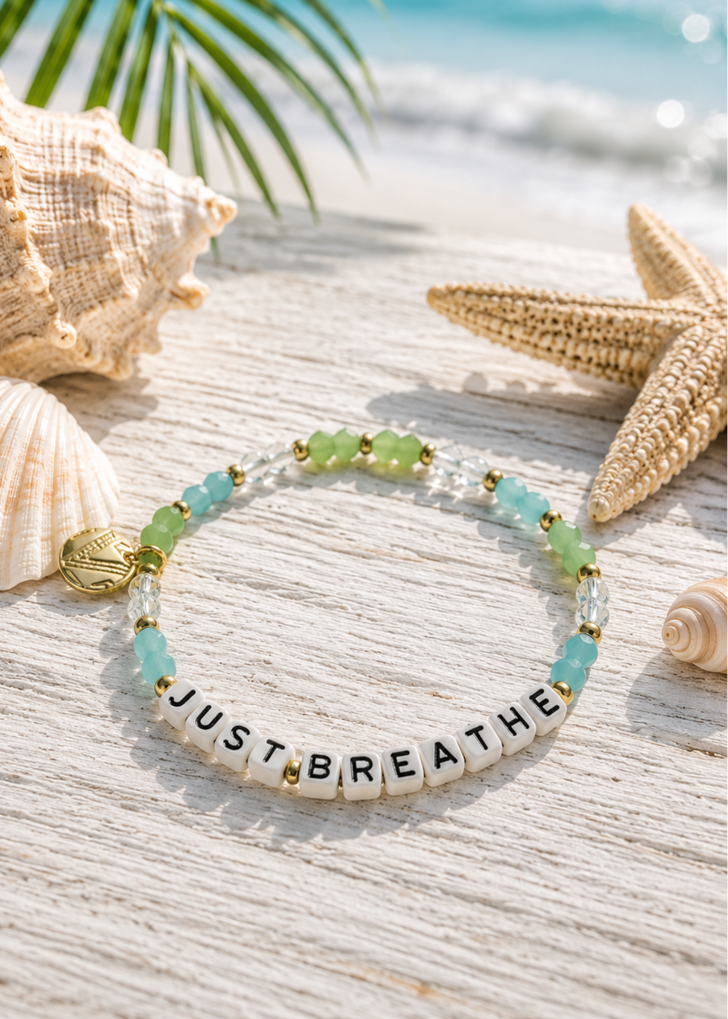 A beaded stretch bracelet with letter beads spelling 'JUST BREATHE' lies on a white wooden surface styled with seashells, a starfish, and a tropical beach background.