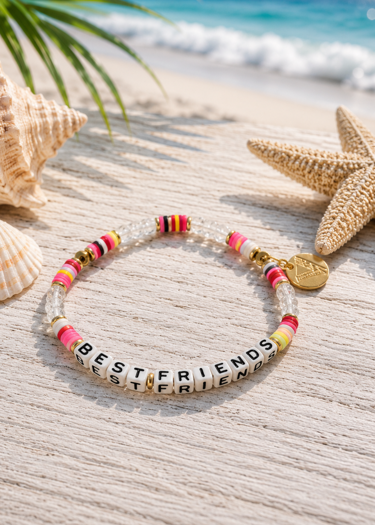 A beaded bracelet with clear acrylic beads spelling 'BEST FRIENDS' in black letters, pink and yellow striped beads, gold spacer beads, and a small gold coin charm, displayed on a sandy beach with seashells and a starfish.