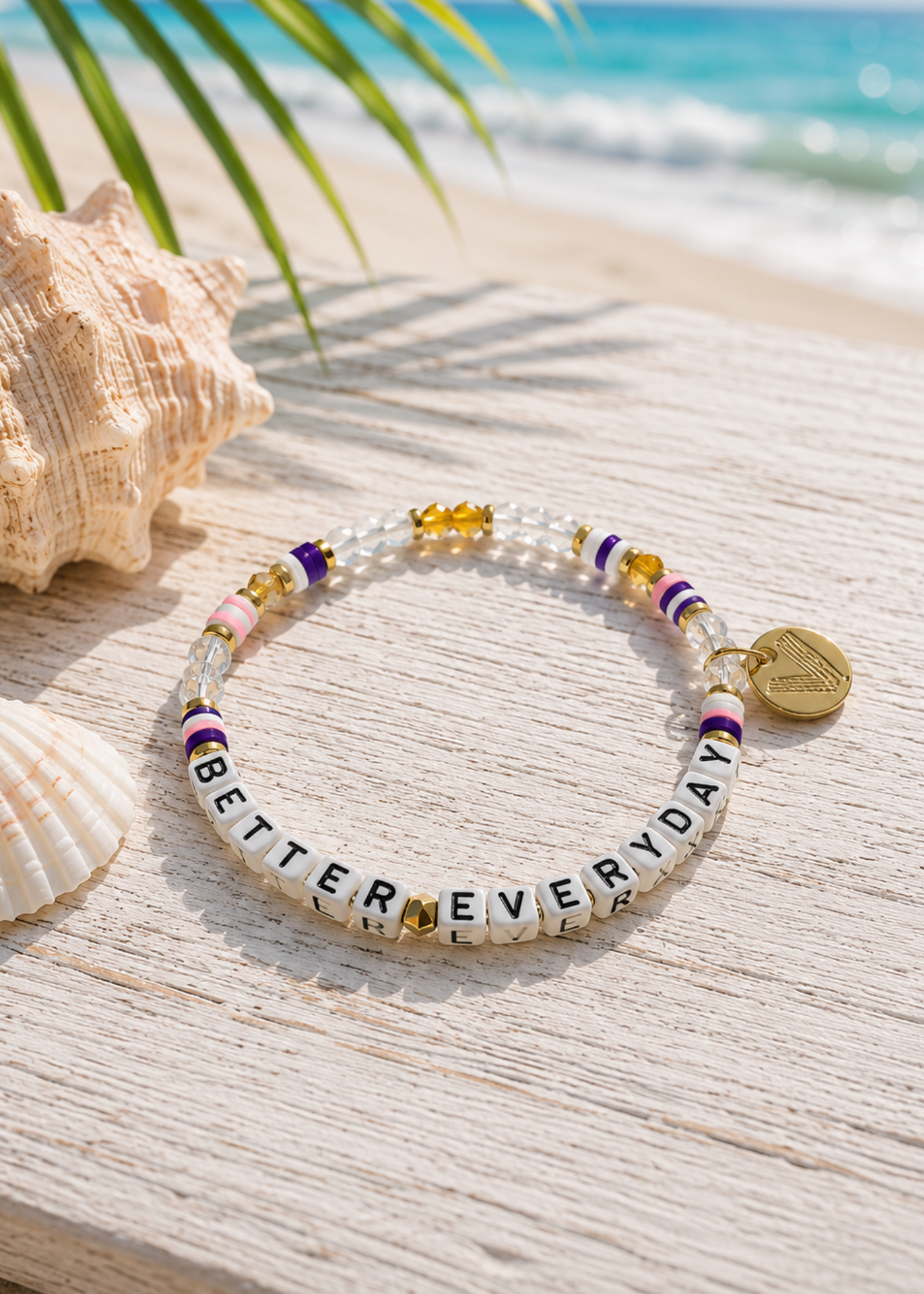 A beaded stretch bracelet displayed on a light wood surface with seashells and a beach background. The bracelet features white letter beads spelling 'BETTER EVERYDAY', purple and gold spacer beads, and a small gold heart charm.