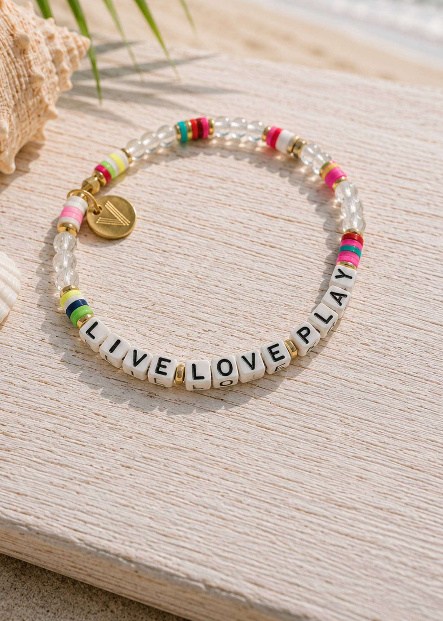 A beaded stretch bracelet displayed on a light wooden surface with a seashell prop. The bracelet features clear faceted beads, colorful spacer beads in pink, green, yellow, and blue, white letter tiles spelling 'LIVE LOVE PLAY', and a small gold charm.