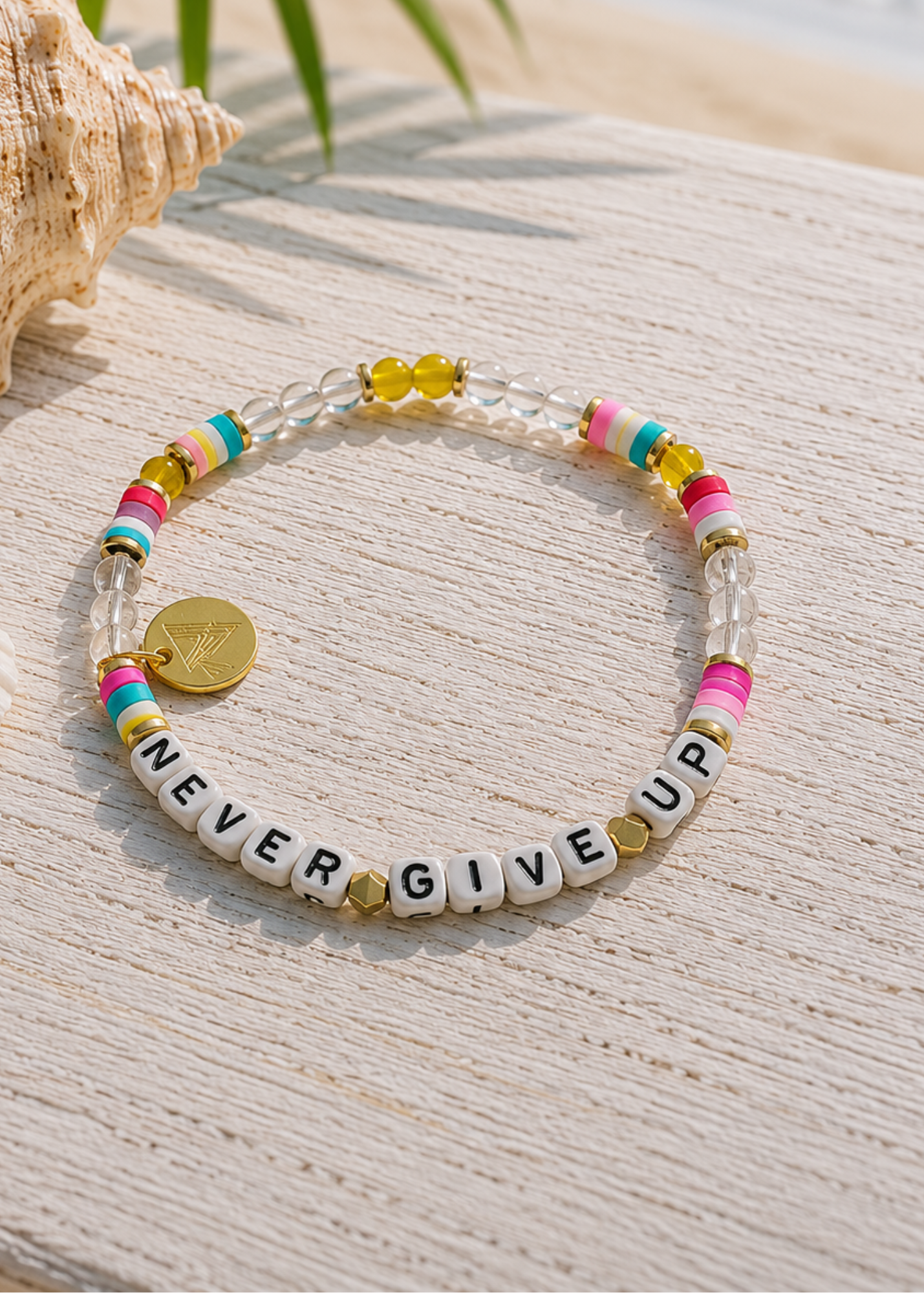A beaded stretch bracelet with white letter beads spelling 'NEVER GIVE UP', clear round beads, small colorful accent beads, and a gold coin charm, displayed on a light wood surface.