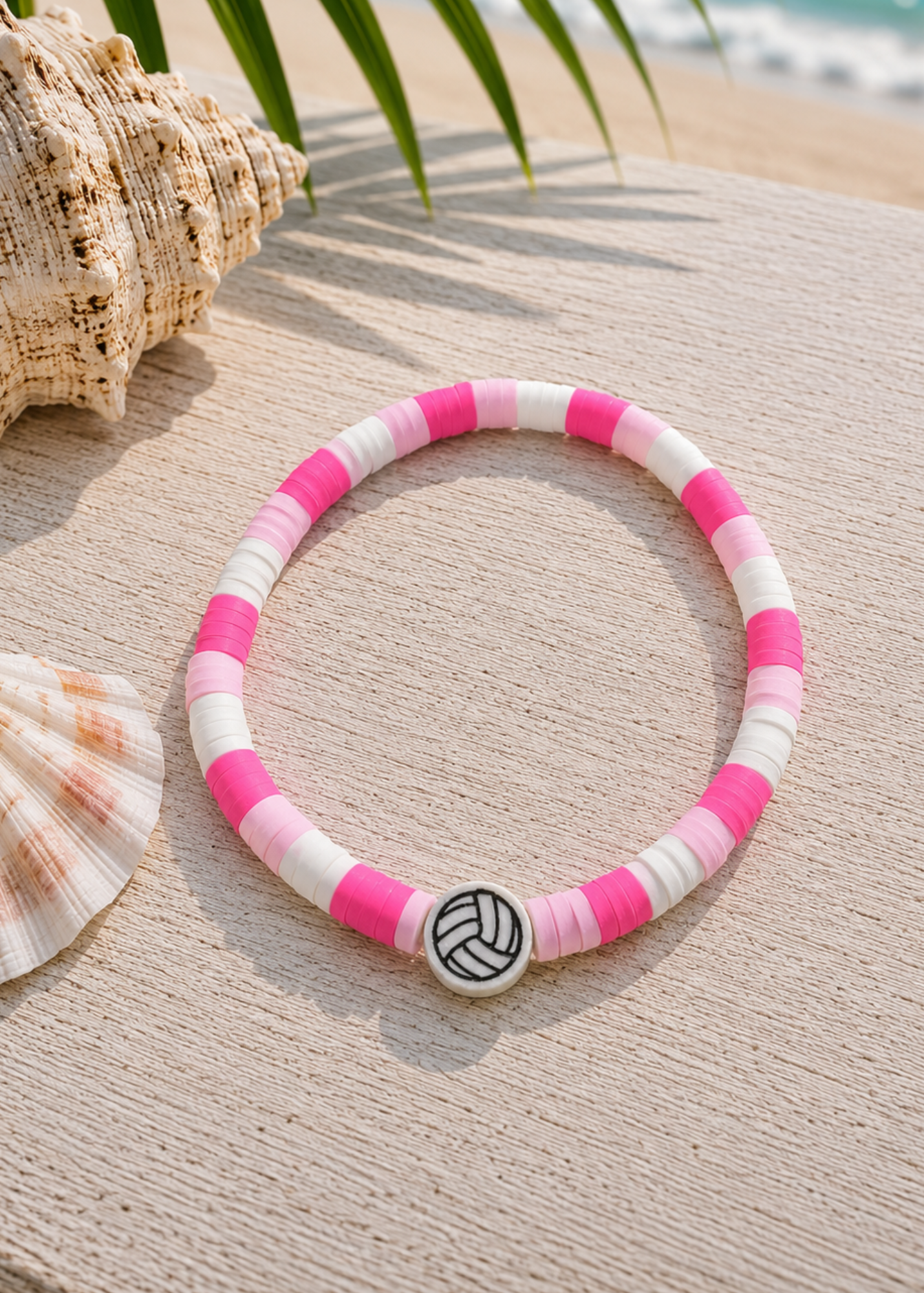 A beaded bracelet with alternating pink and white beads, featuring a central silver-toned charm shaped like a volleyball, displayed on a sandy surface with seashells and palm leaves.