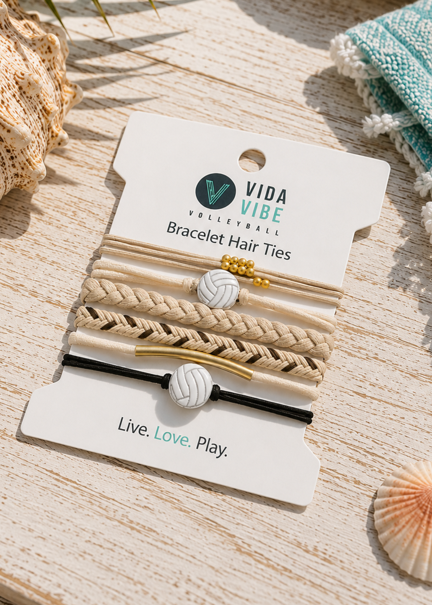 Vida Vibe Volleyball bracelet hair ties on a white display card, styled on a light wood surface with seashells and a woven textile.