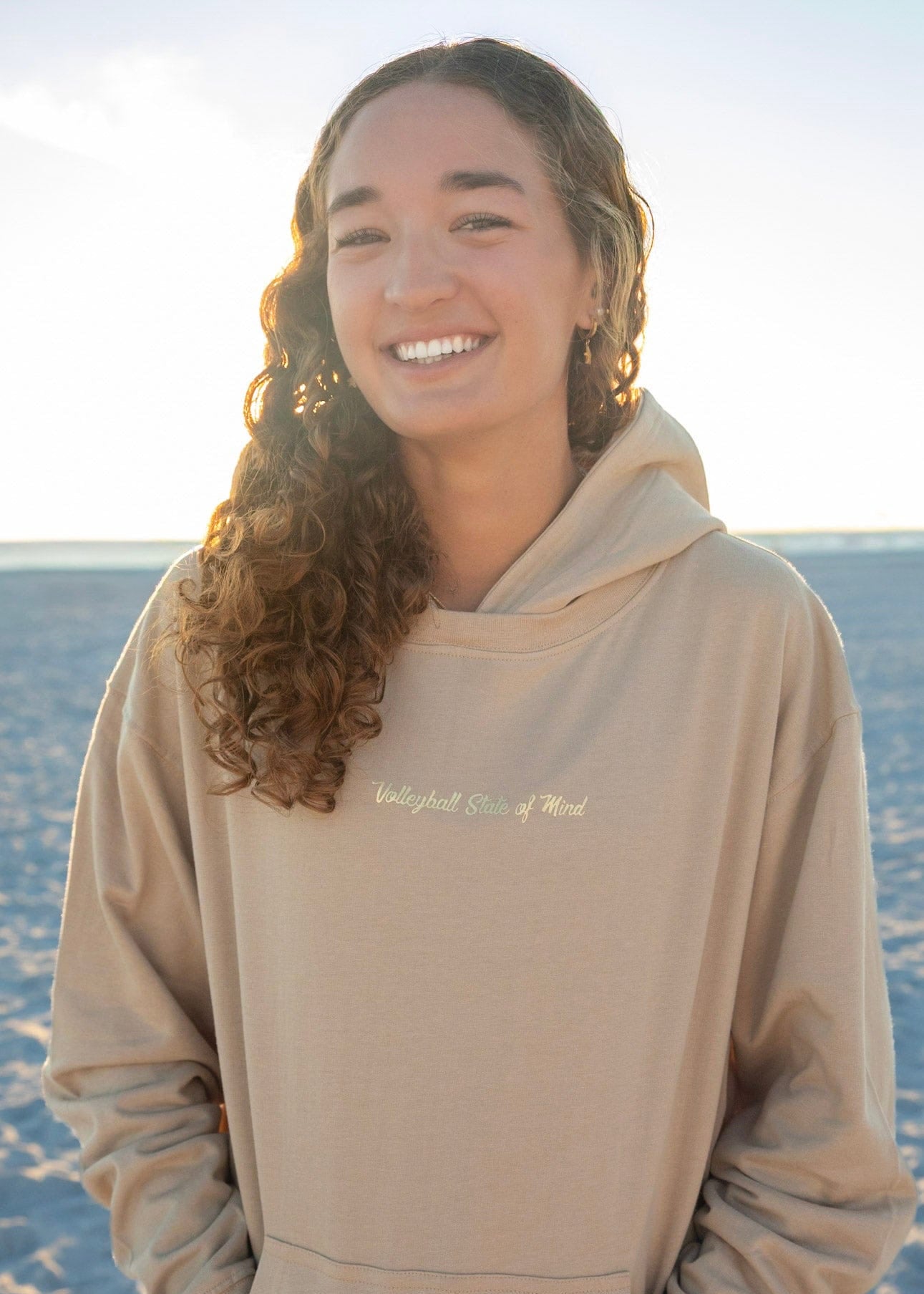 Woman wearing a beige hoodie with text on it, standing on a beach.
