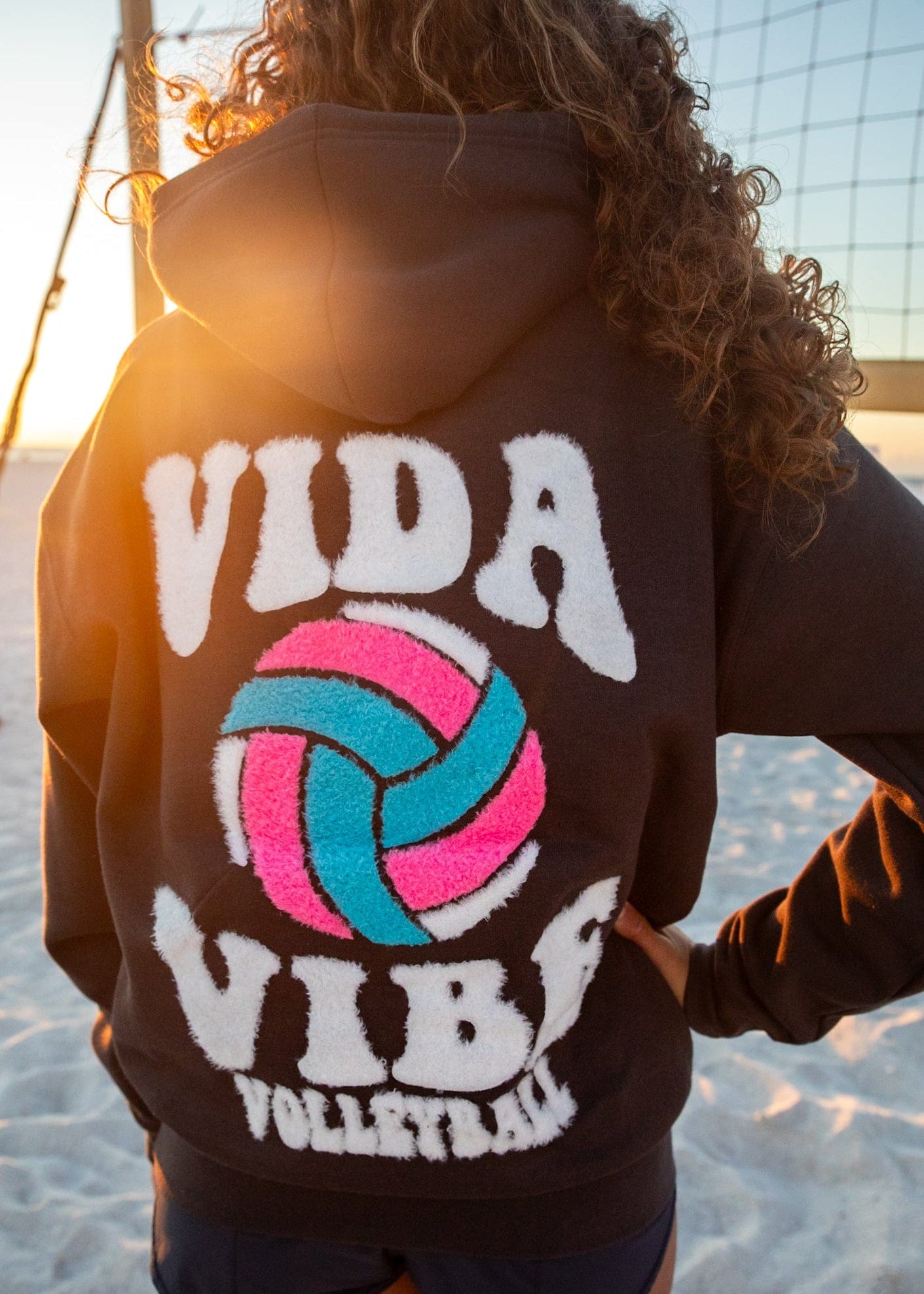Person wearing a hoodie with 'Vida Vibe Volleyball' design on a beach.