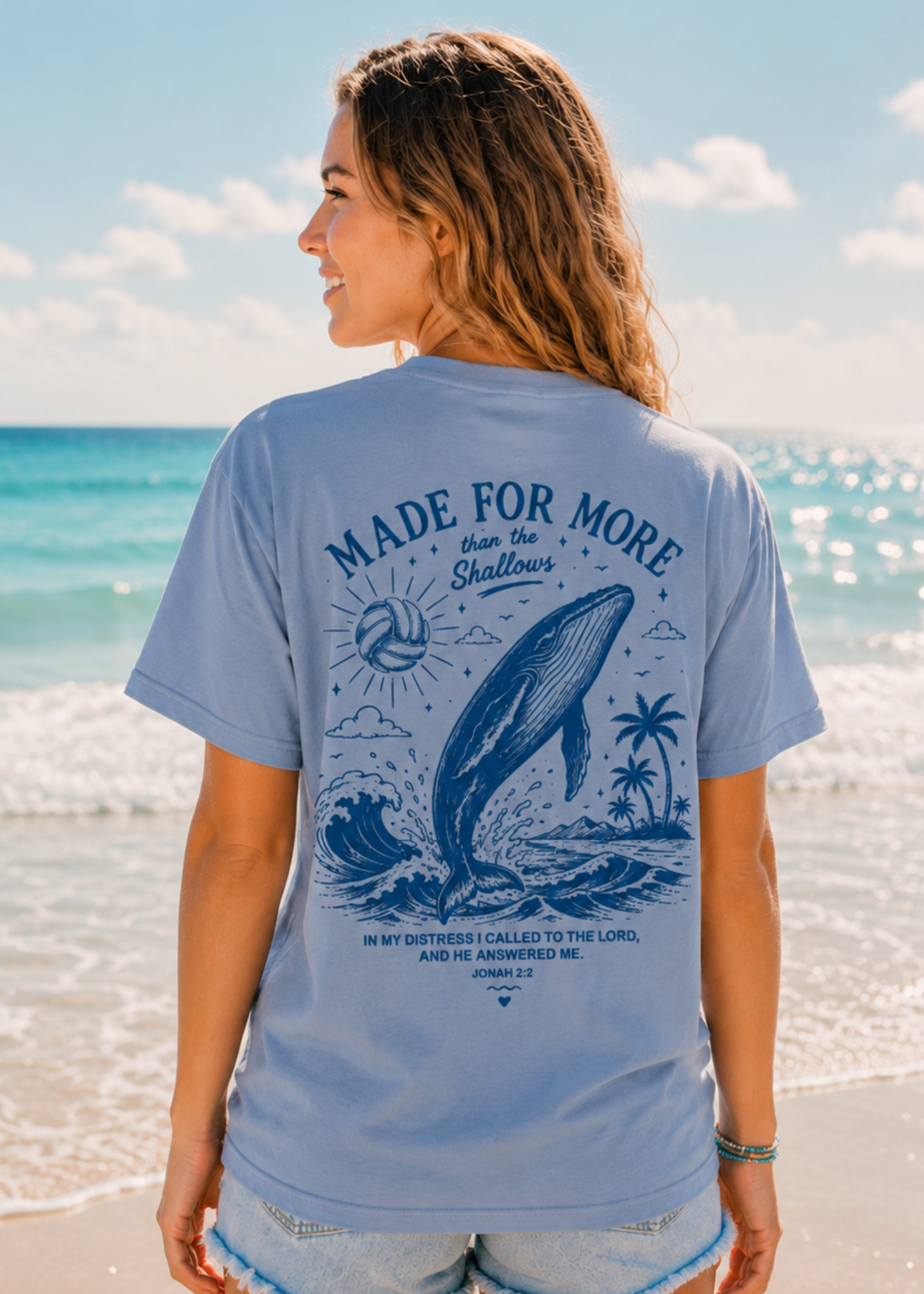 A woman standing on a beach wearing a light blue graphic volleyball t-shirt with a navy blue whale and scripture print on the back.