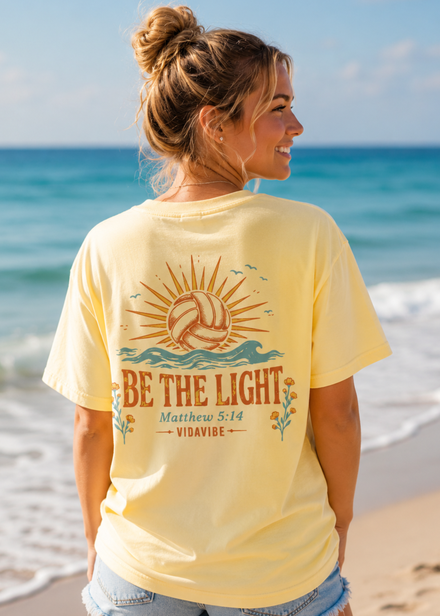 A woman standing on a beach wearing a pale yellow graphic T-shirt with a volleyball and sun design on the back, plus the text 'BE THE LIGHT Matthew 5:14 VIDAYBE'.