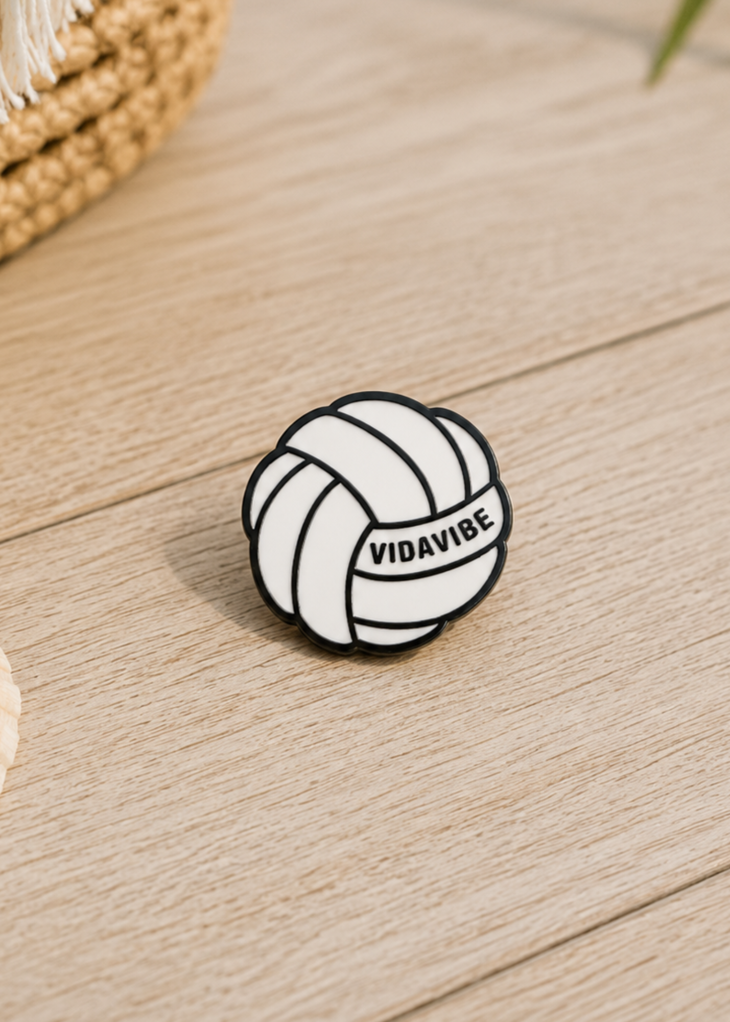 A small volleyball-themed enamel pin with the text 'VIDAVIBE' printed on it, resting on a light wood surface.