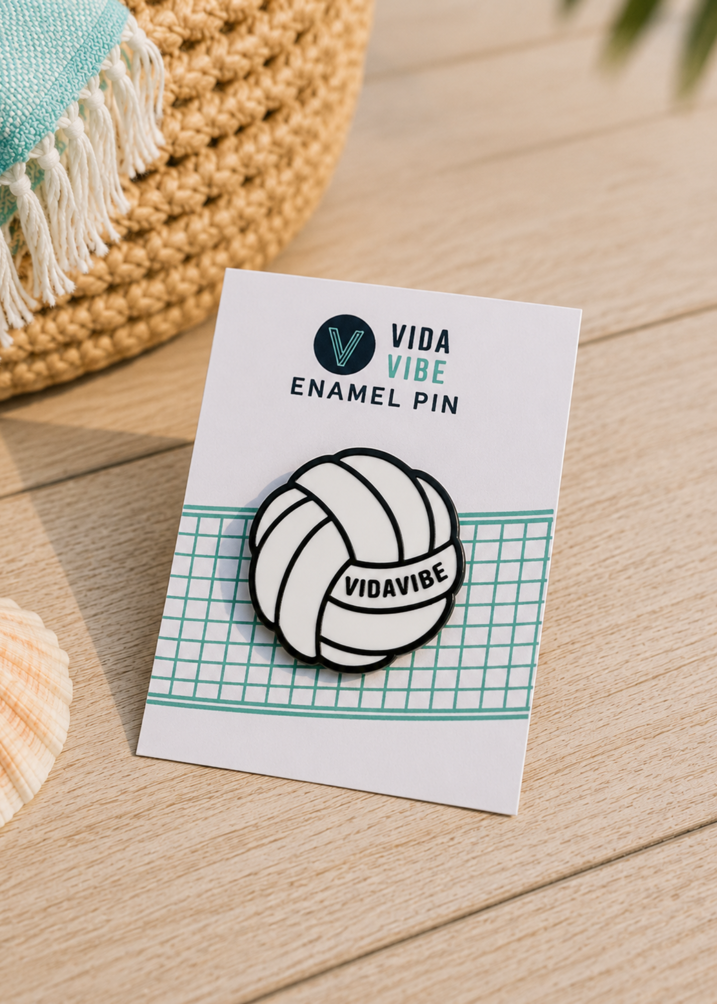 A black and white enamel pin shaped like a volleyball with the word 'VIDAVIBE' on it, mounted on a white display card that reads 'VIDA VIBE ENAMEL PIN'. The card is placed on a light wood surface next to a woven basket and a seashell.