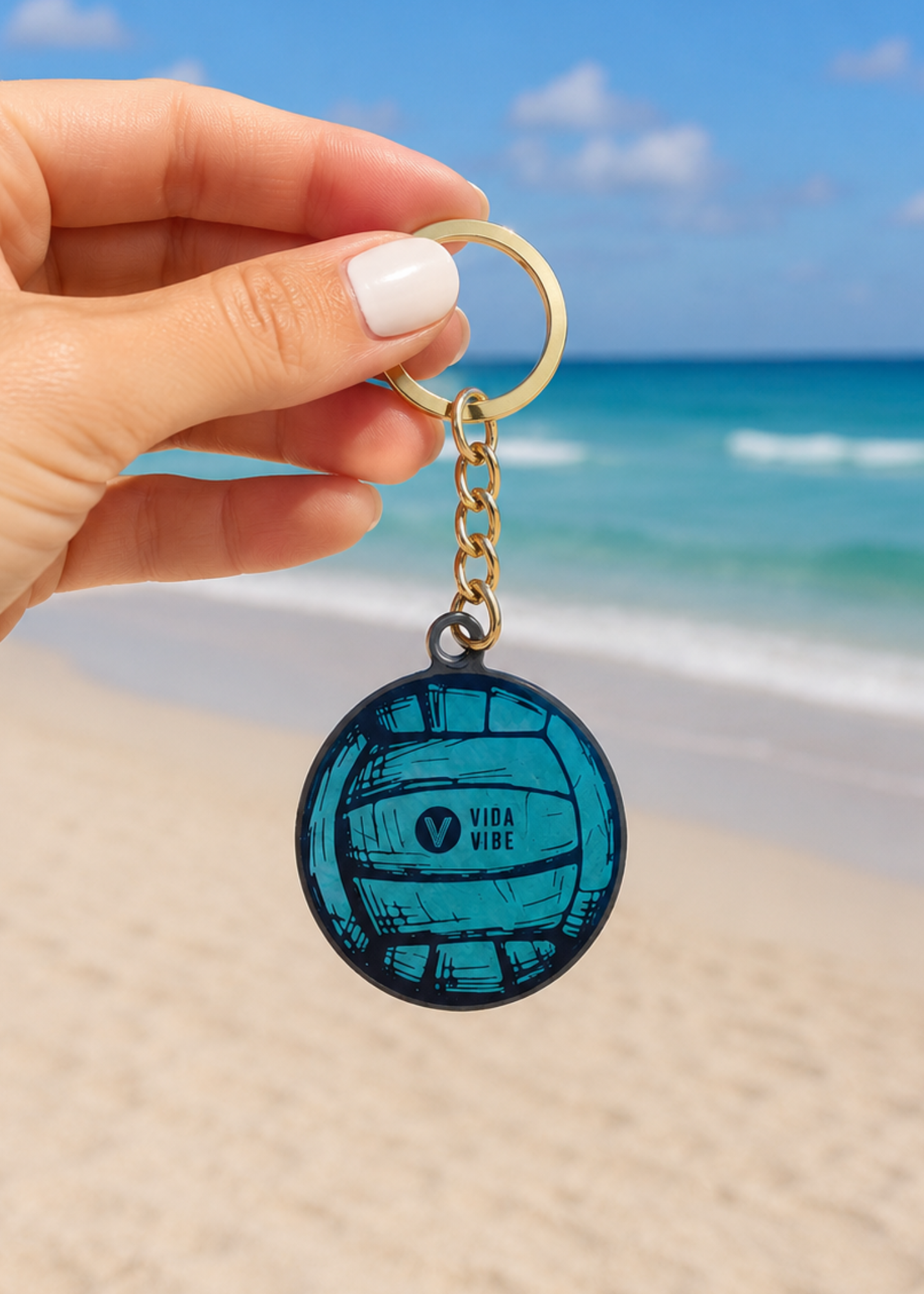 A hand with white nail polish holds a round keychain featuring a teal and black volleyball design with the text 'VIDA VIDE' on a gold split ring, set against a blurred beach and ocean background.