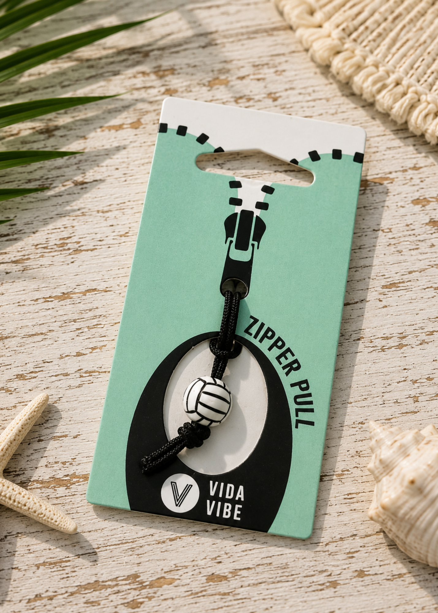 A zipper pull charm with a black cord, metal zipper pull, and white-and-black striped bead, attached to a mint and black retail card labeled 'ZIPPER PULL' and 'VIDA VIBE', photographed on a whitewashed wood surface with seashell and palm leaf props.