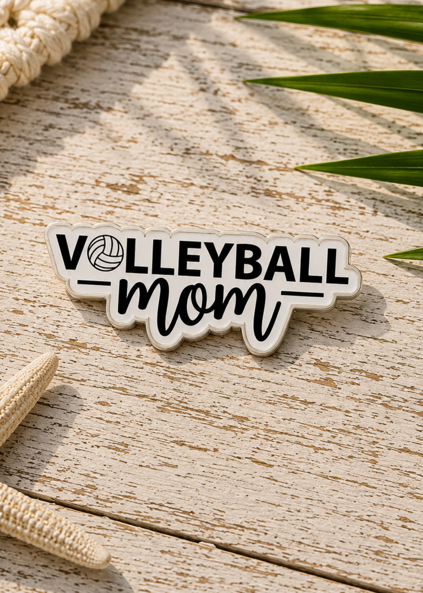 A die-cut enamel pin reading 'VOLLEYBALL mom' with a volleyball icon, placed on a weathered wood surface with beach-themed props like a rope, starfish, and green leaves.
