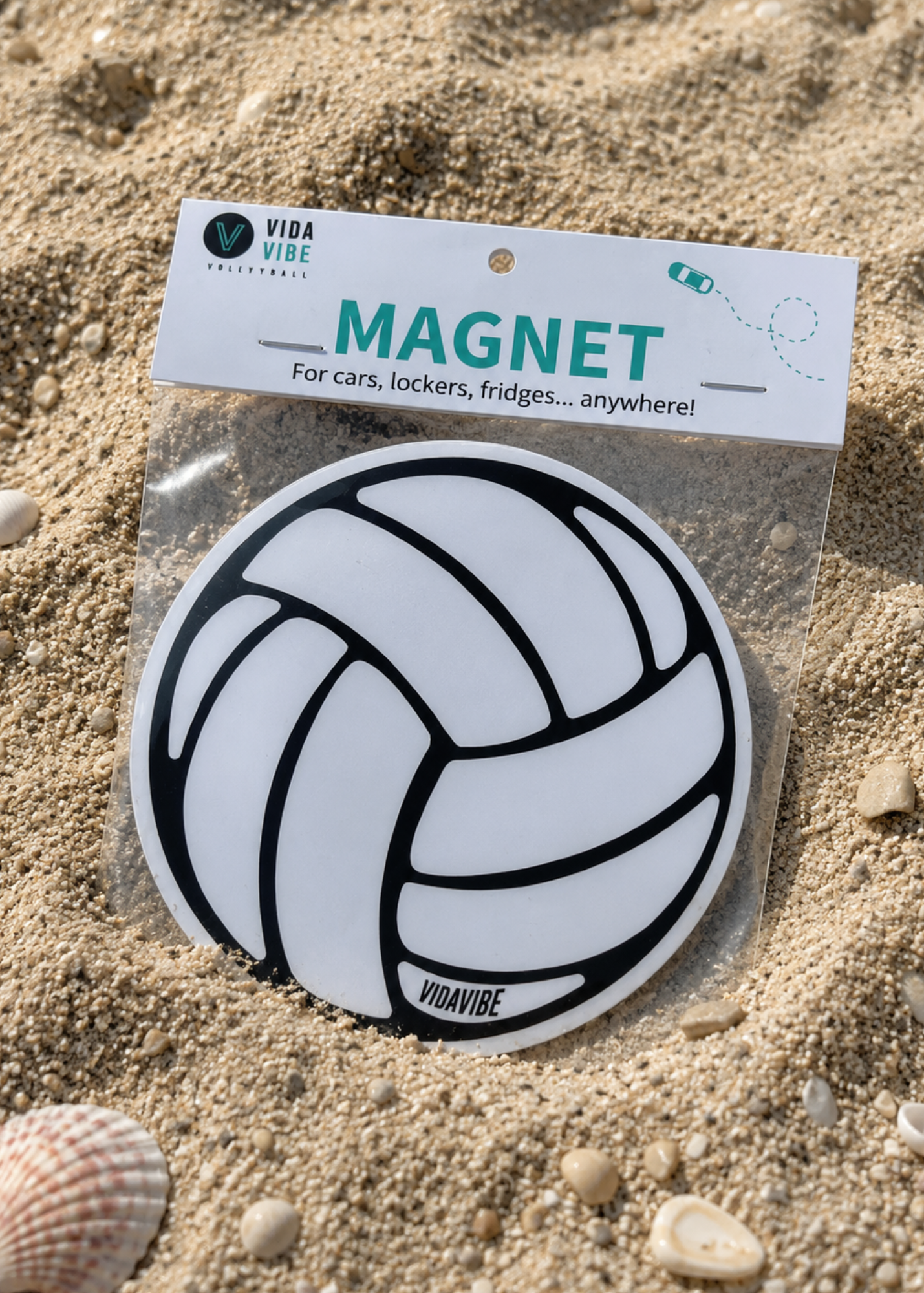 A round volleyball-themed magnet in its clear plastic packaging with a white header card, placed on a sandy beach with small seashells nearby.