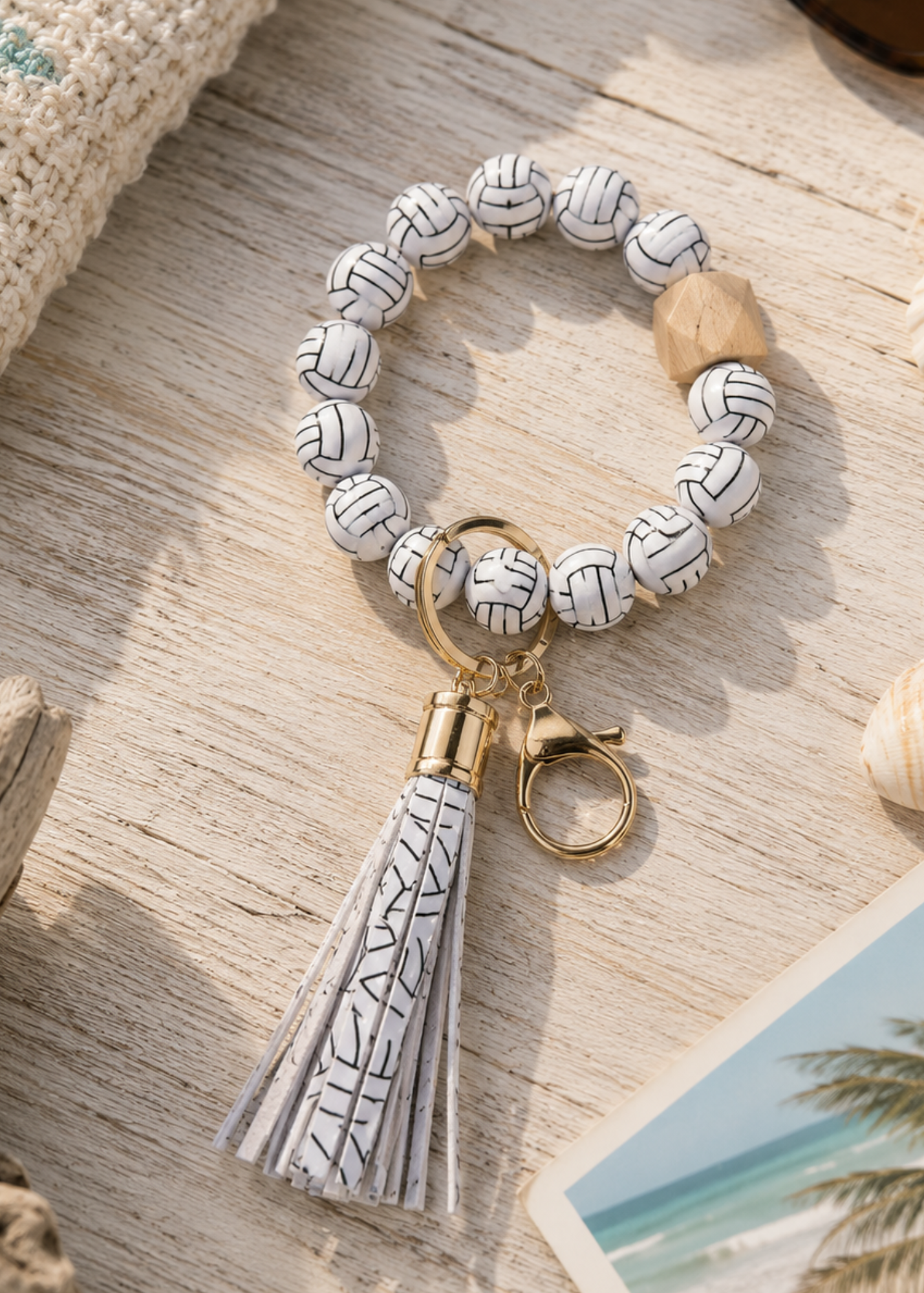 A beaded keychain featuring white volleyball-patterned beads, a gold-tone split ring, a white tassel with black line art, and a small wooden bead, displayed on a light wood surface.