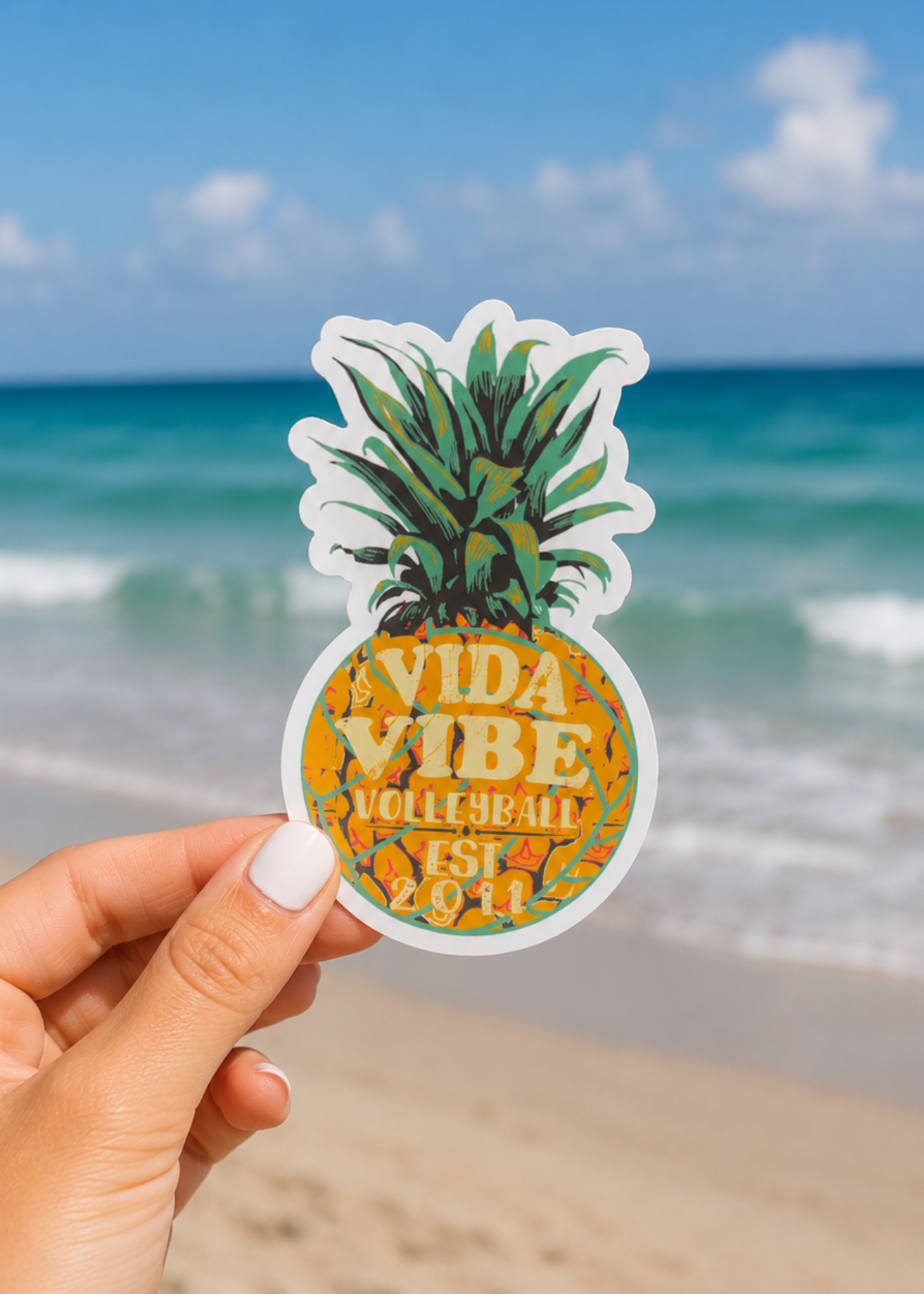 A hand holds a die-cut pineapple sticker with the text 'VIDA VIBE VOLLEYBALL EST. 2011' against a blurred beach and ocean background.