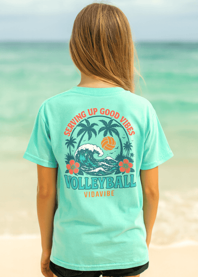 Person wearing a teal t-shirt with volleyball design and text on a beach.