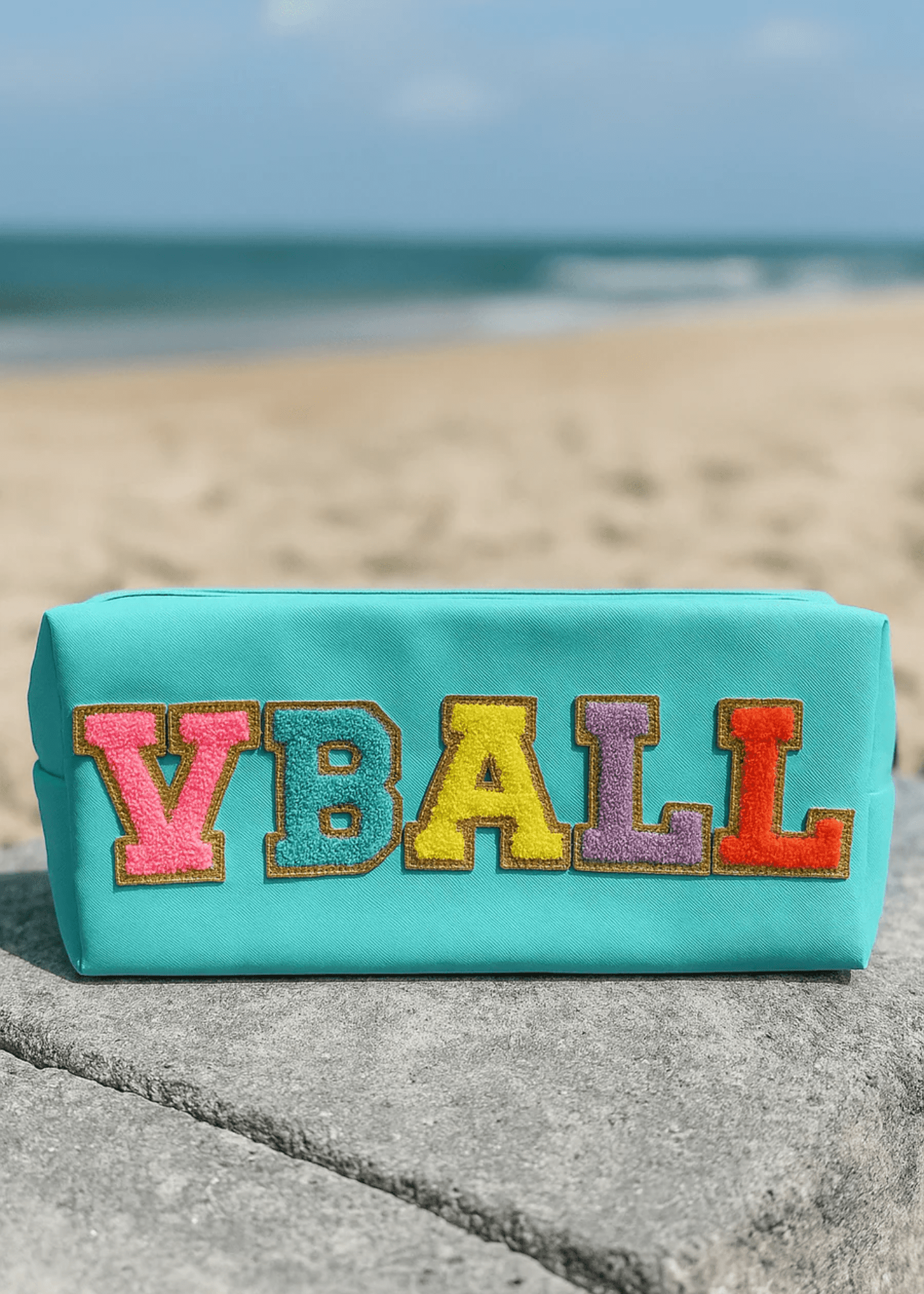 Turquoise pouch with colorful 'VBALL' letters on a beach.